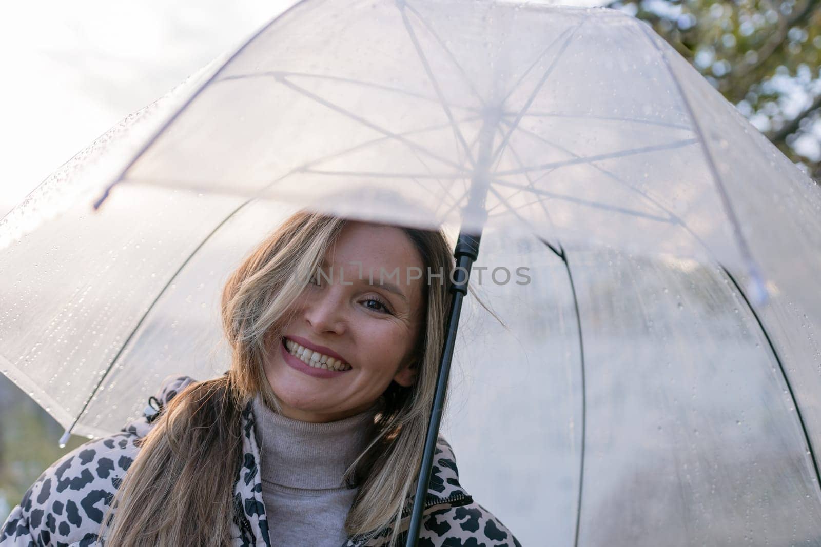 A woman is smiling under a clear umbrella. The umbrella is white and has a black handle. The woman is wearing a jacket and a scarf