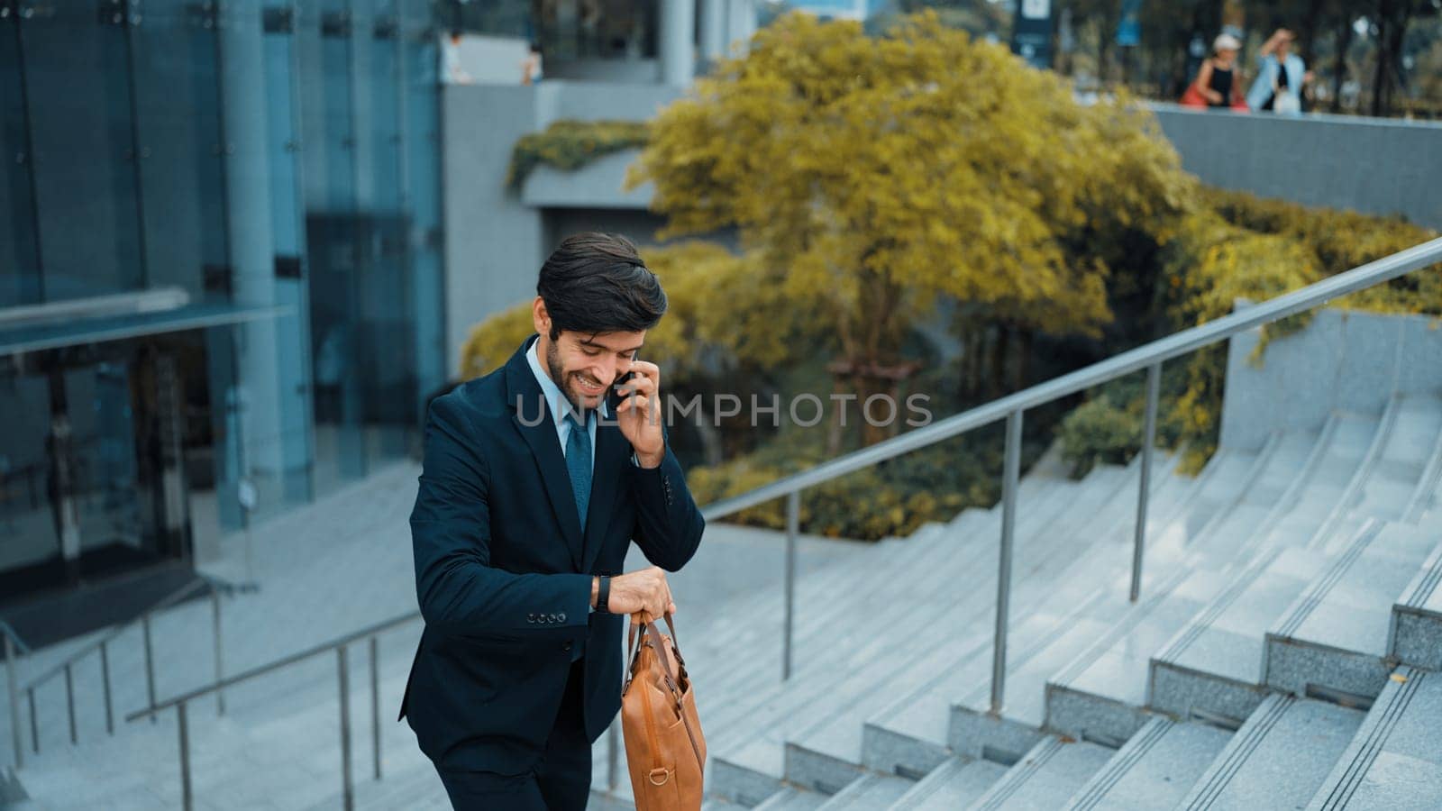 Smiling handsome business man looking at time and walking up stair at park while calling manager about startup project business idea. Investor talking to colleague while going to work. Exultant.