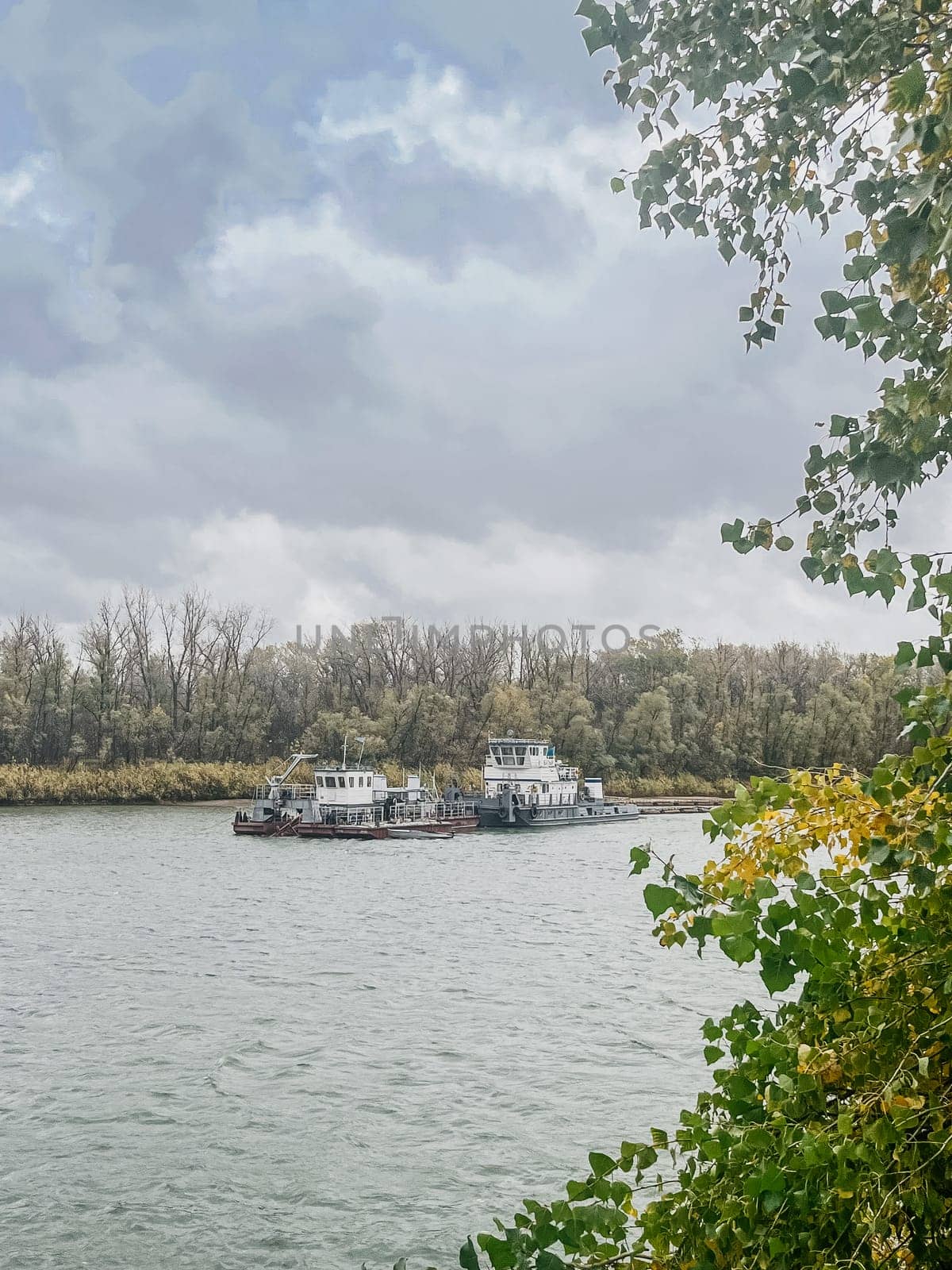 The image captures the serene scene of tugboats navigating the river under a cloudy sky with autumn trees on the shore.