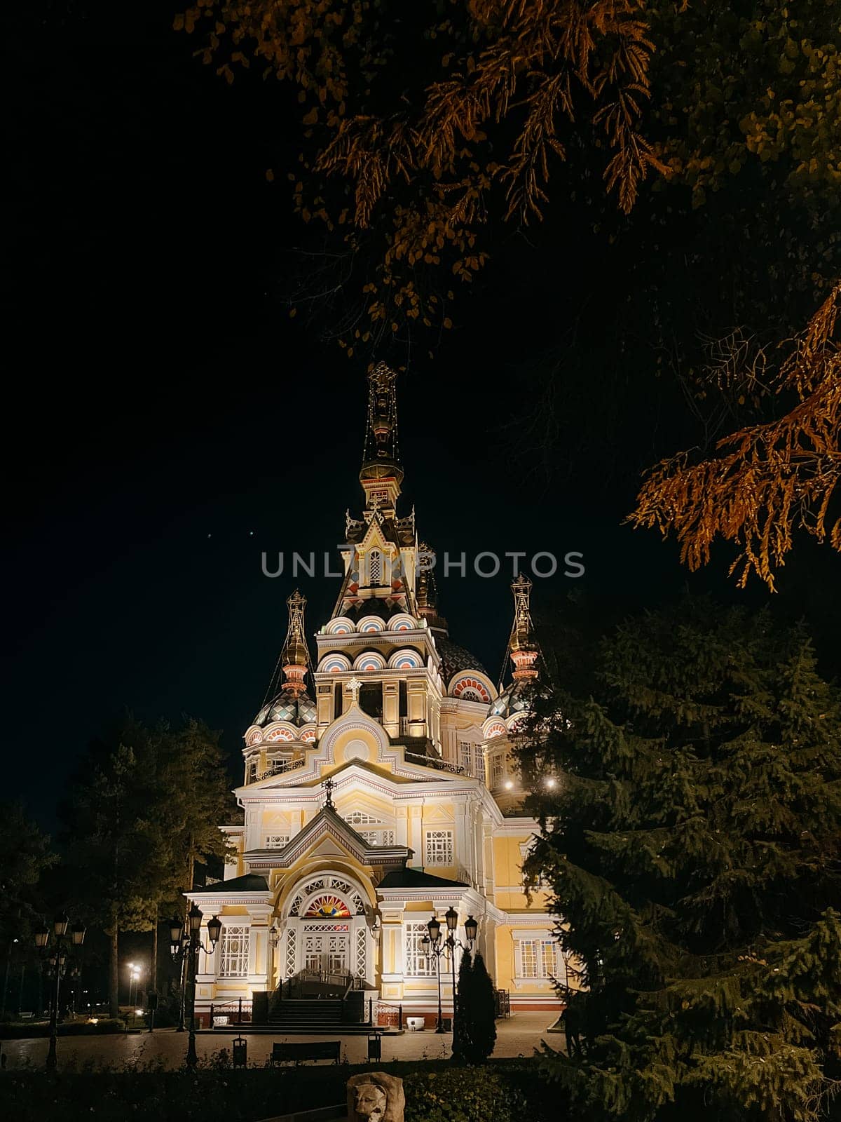 A stunning Russian Orthodox church illuminated at night, with a vibrant yellow exterior, towering steeple, surrounded by trees and park.