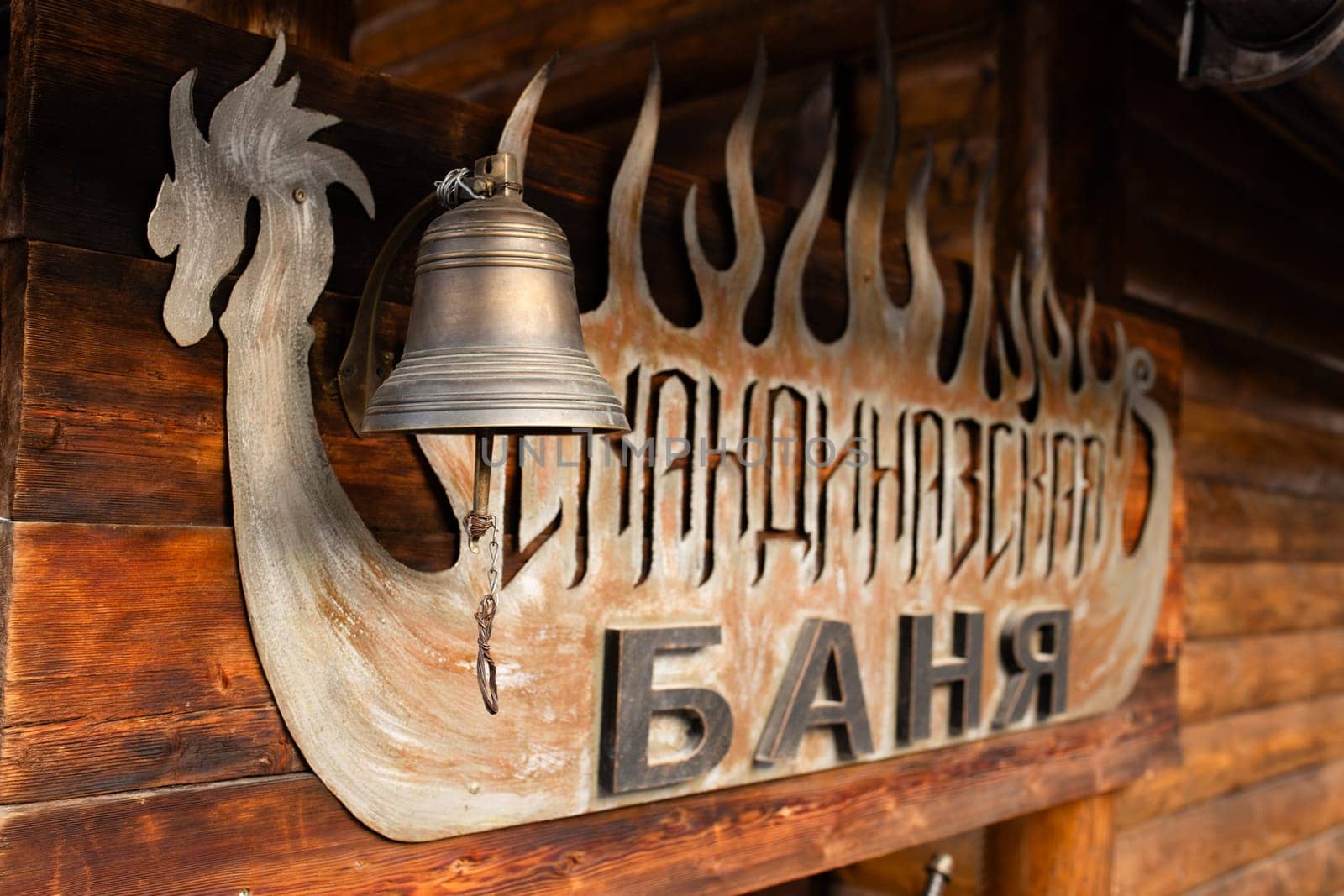 Rustic bell hanging from wooden structure with Banya sign in Cyrillic letters by Pukhovskiy