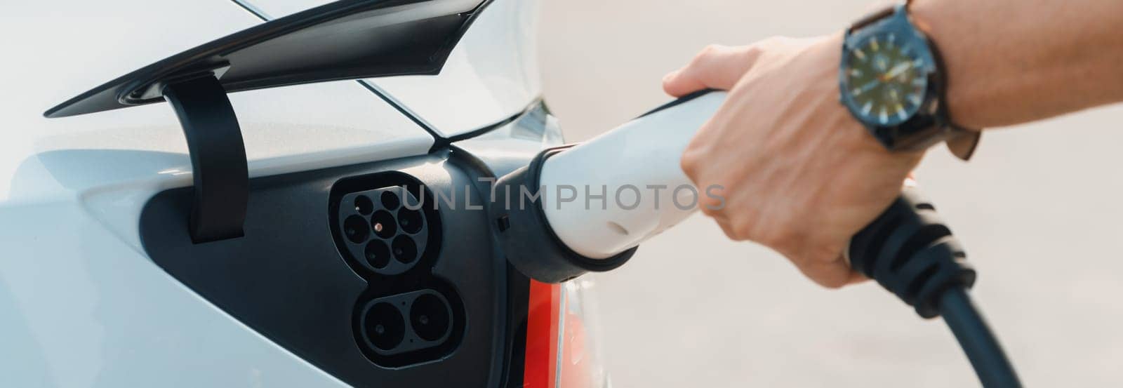 Closeup hand put EV charger to recharge electric car's battery in city commercial parking lot. Rechargeable EV car for sustainable environmental friendly urban travel. Panorama Expedient