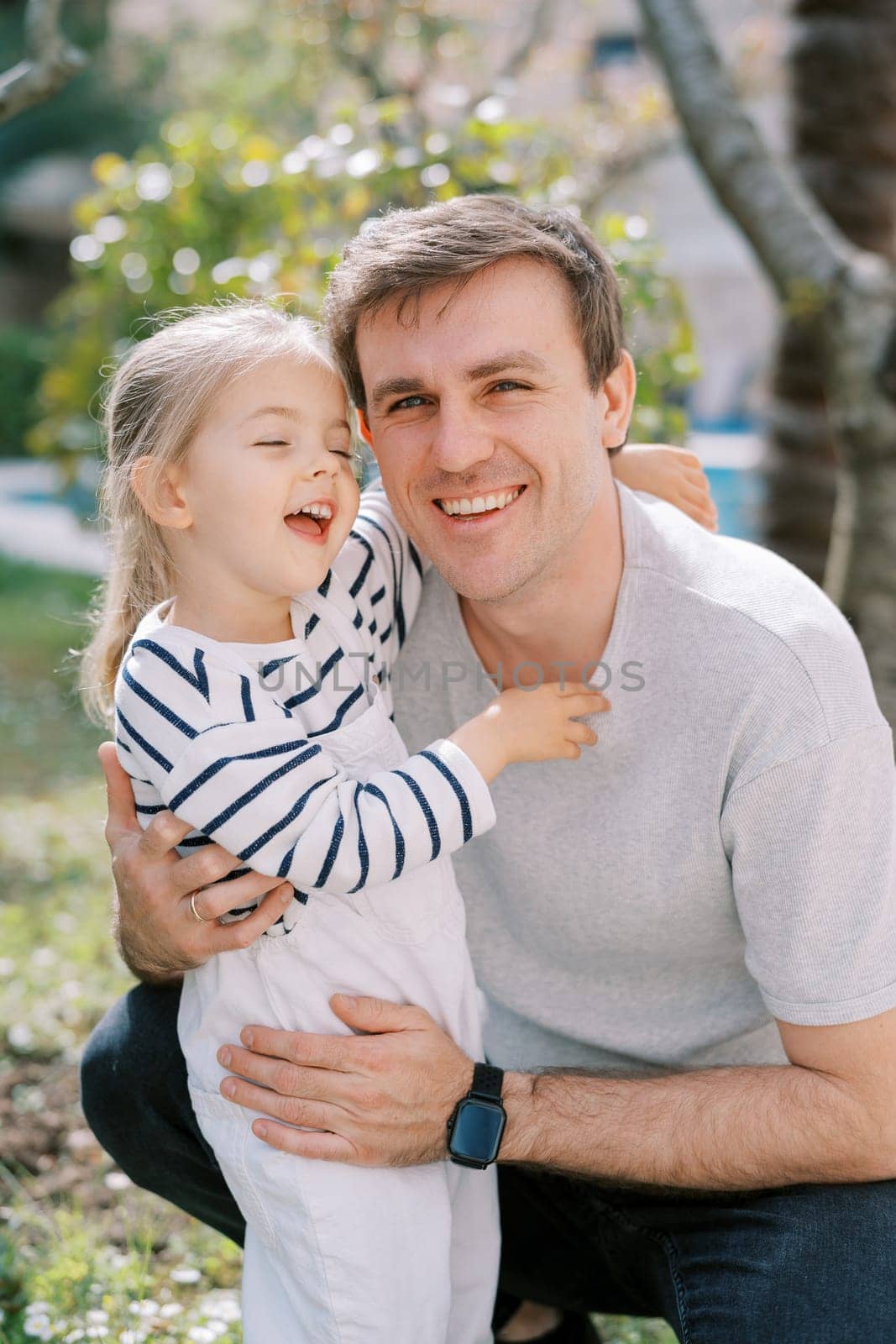 Little laughing girl hugs the neck of her smiling dad squatting in the garden. High quality photo
