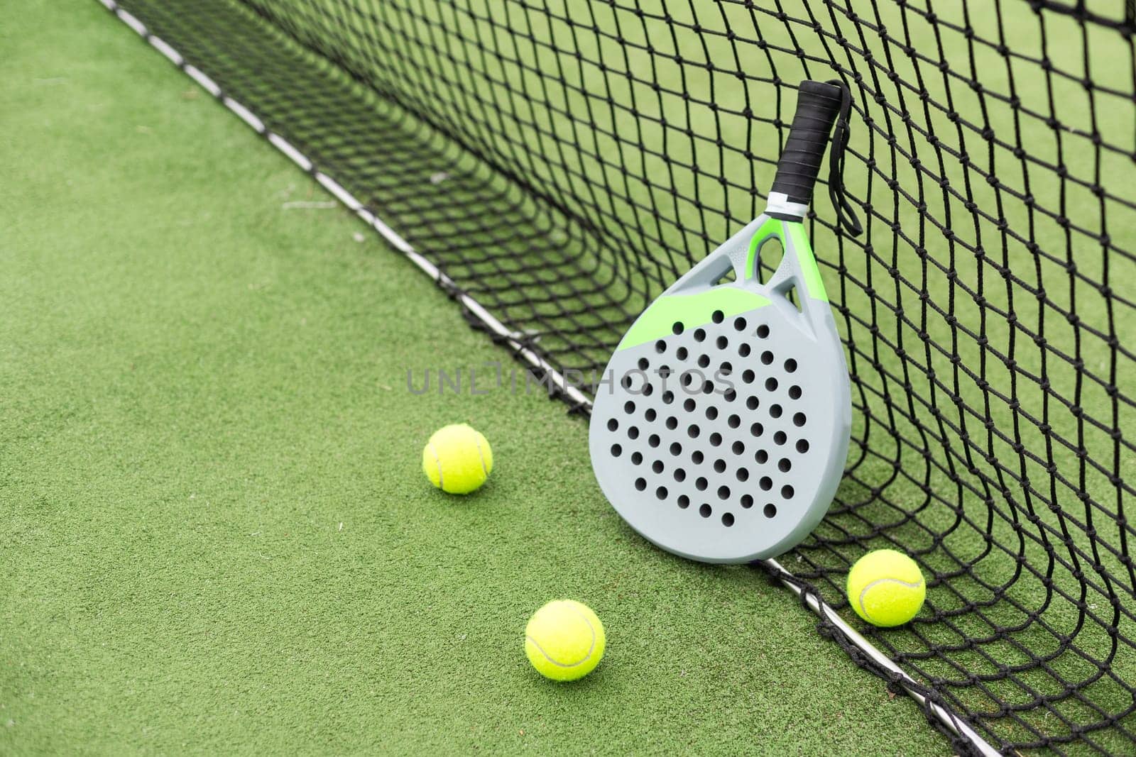 Yellow balls on grass turf near padel tennis racket behind net in green court outdoors with natural lighting. Paddle is a racquet game. Professional sport concept with copy space. High quality photo