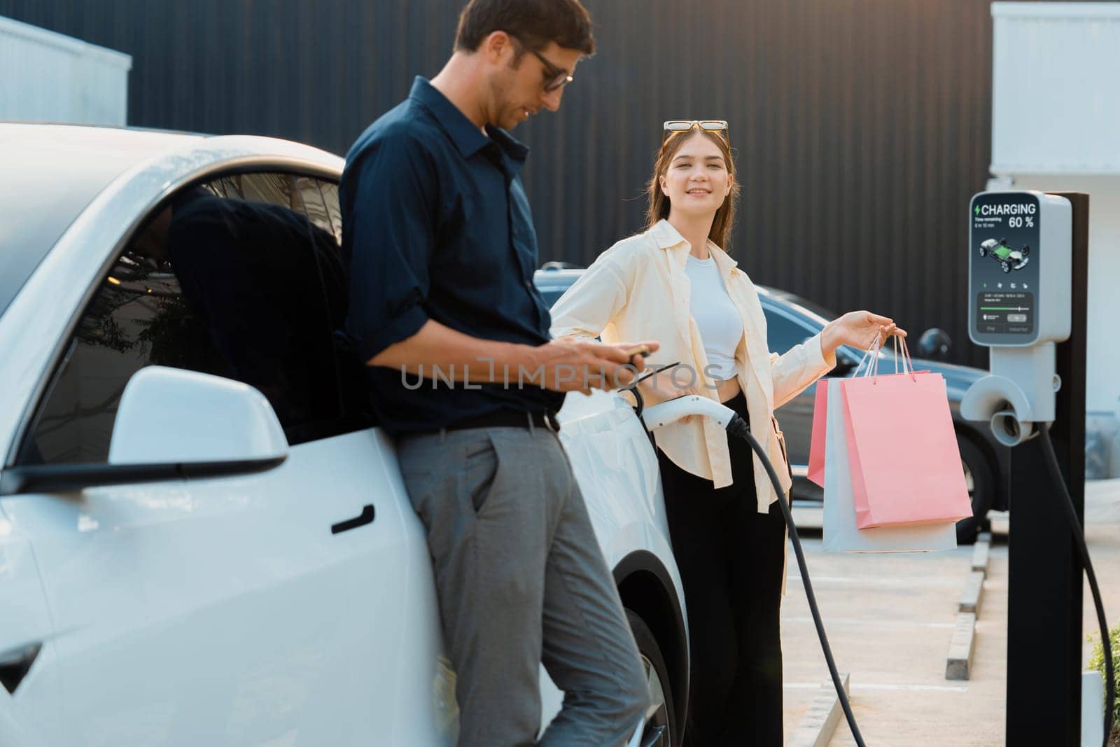 Young couple holding shopping bag and use smartphone to pay for electricity for recharging EV car battery from charging station at city mall parking lot. Happy couple go shopping by eco car. Expedient