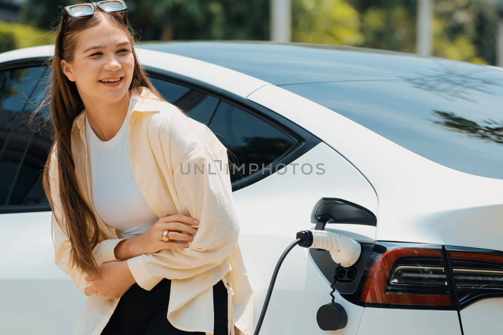 Young woman recharge her EV electric vehicle at green city park parking lot. Urban sustainability lifestyle for environmental friendly EV car with battery charging station. Expedient