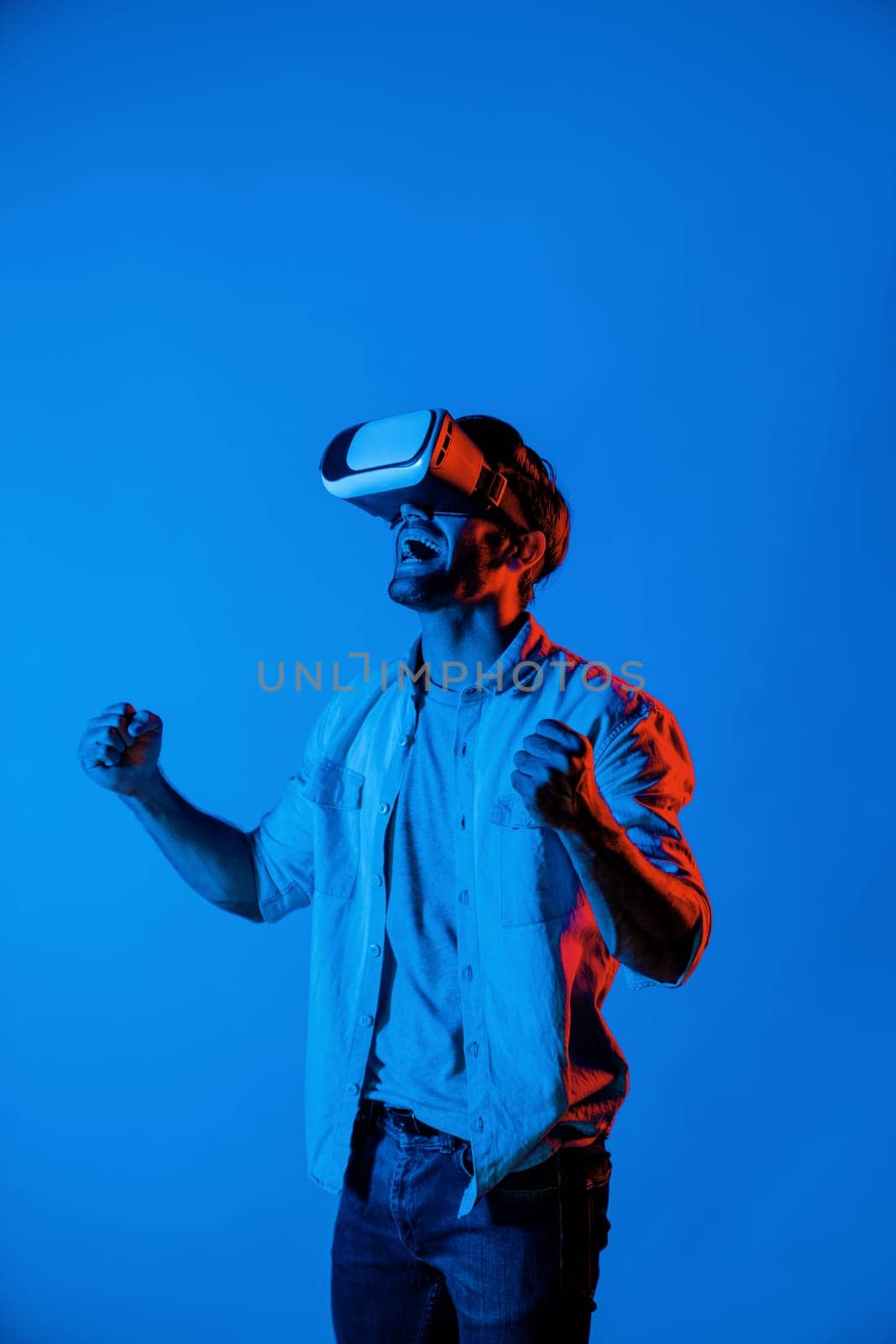 Professional gamer put the hand in the air to celebrate for winning game while wearing VR goggle at colorful neon light background. Caucasian man using technology innovation and enter. Deviation.