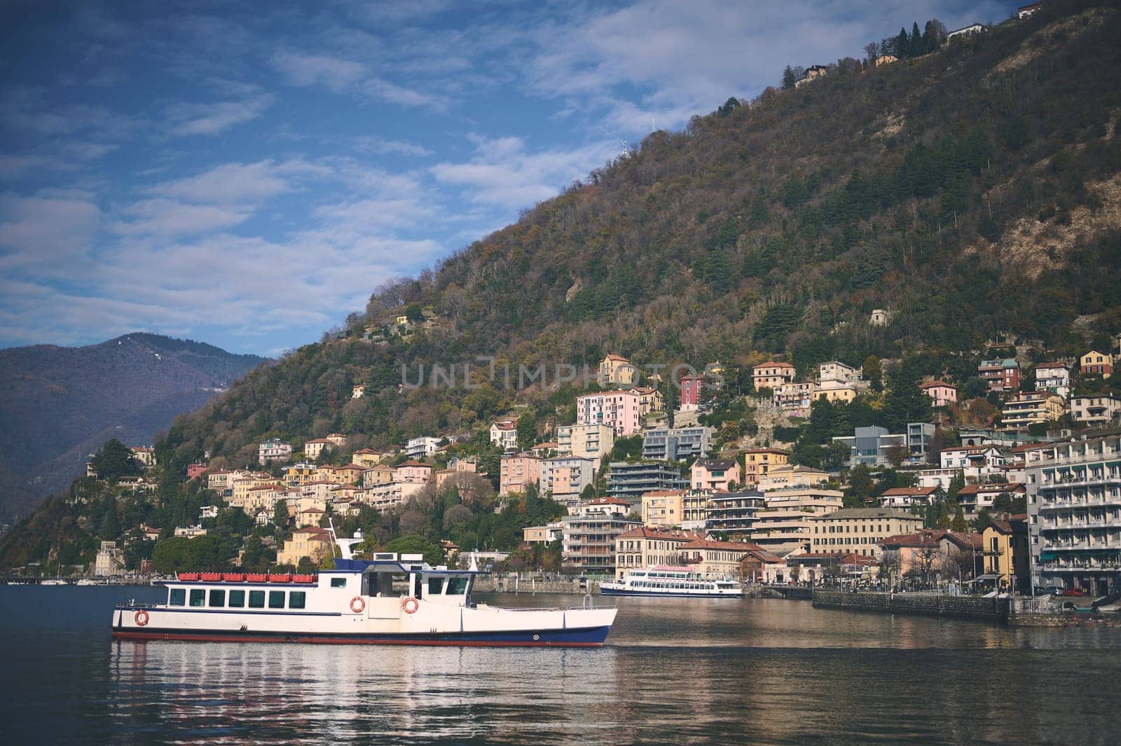 A boat on the lake of Como against blue sky and Italian Alps mountains background. Clear water of the lake reflecting floating boat and houses on the waterfront. Nature background, Tourism and travel.