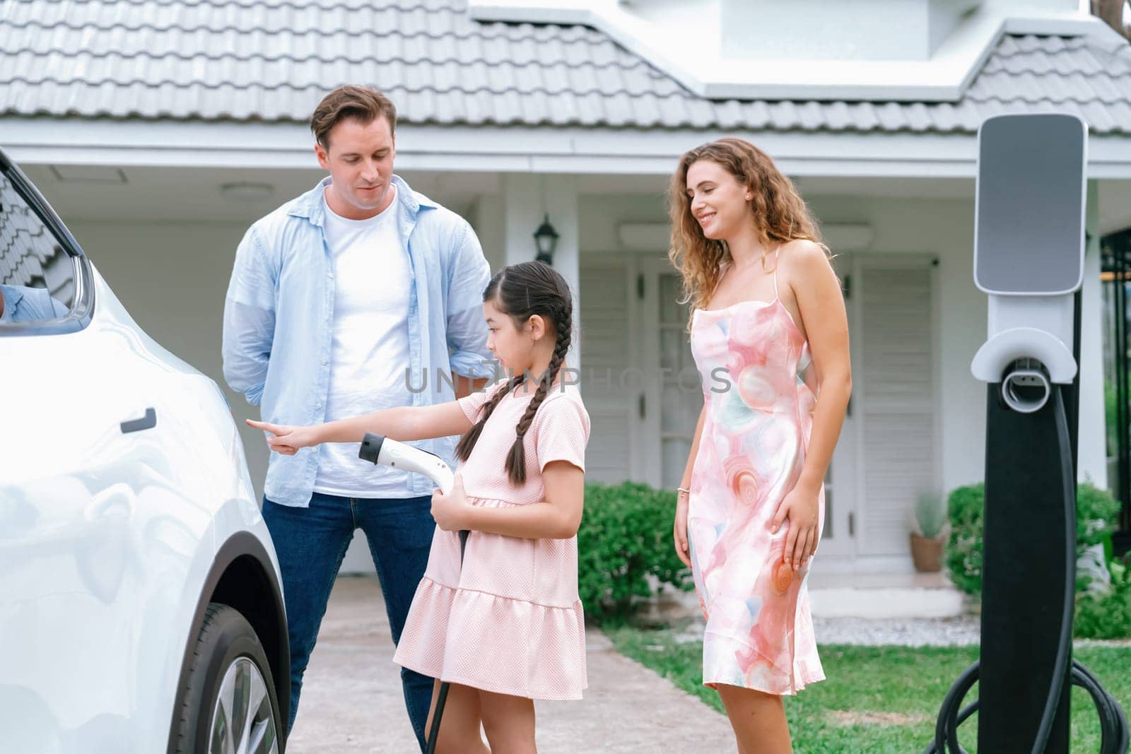 Happy little young girl learn about eco-friendly and energy sustainability as she help her family recharge electric vehicle from home EV charging station. EV car and modern family concept. Synchronos