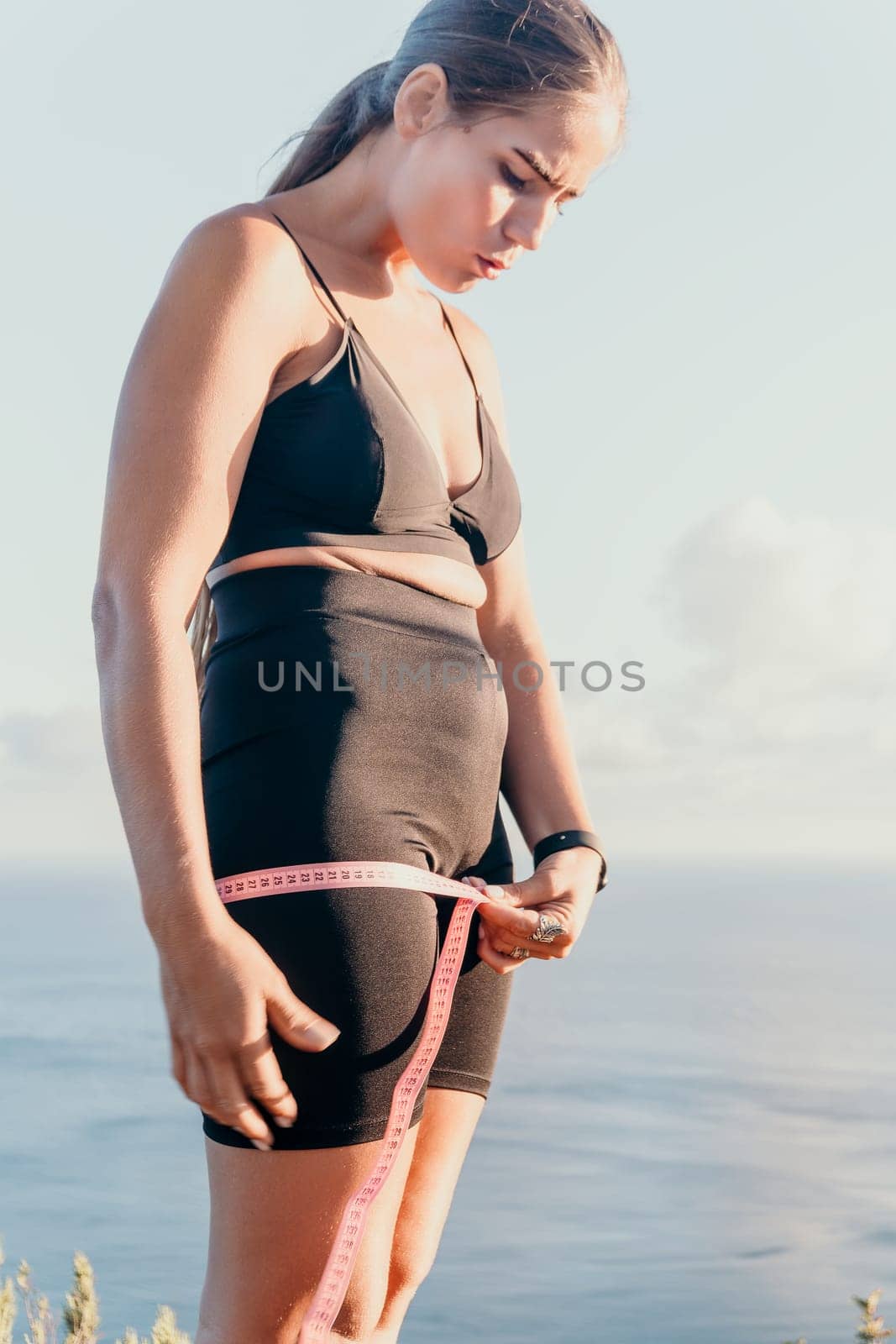 A woman is holding a pink tape measure around her hips. She is wearing black pants and a black top. by panophotograph