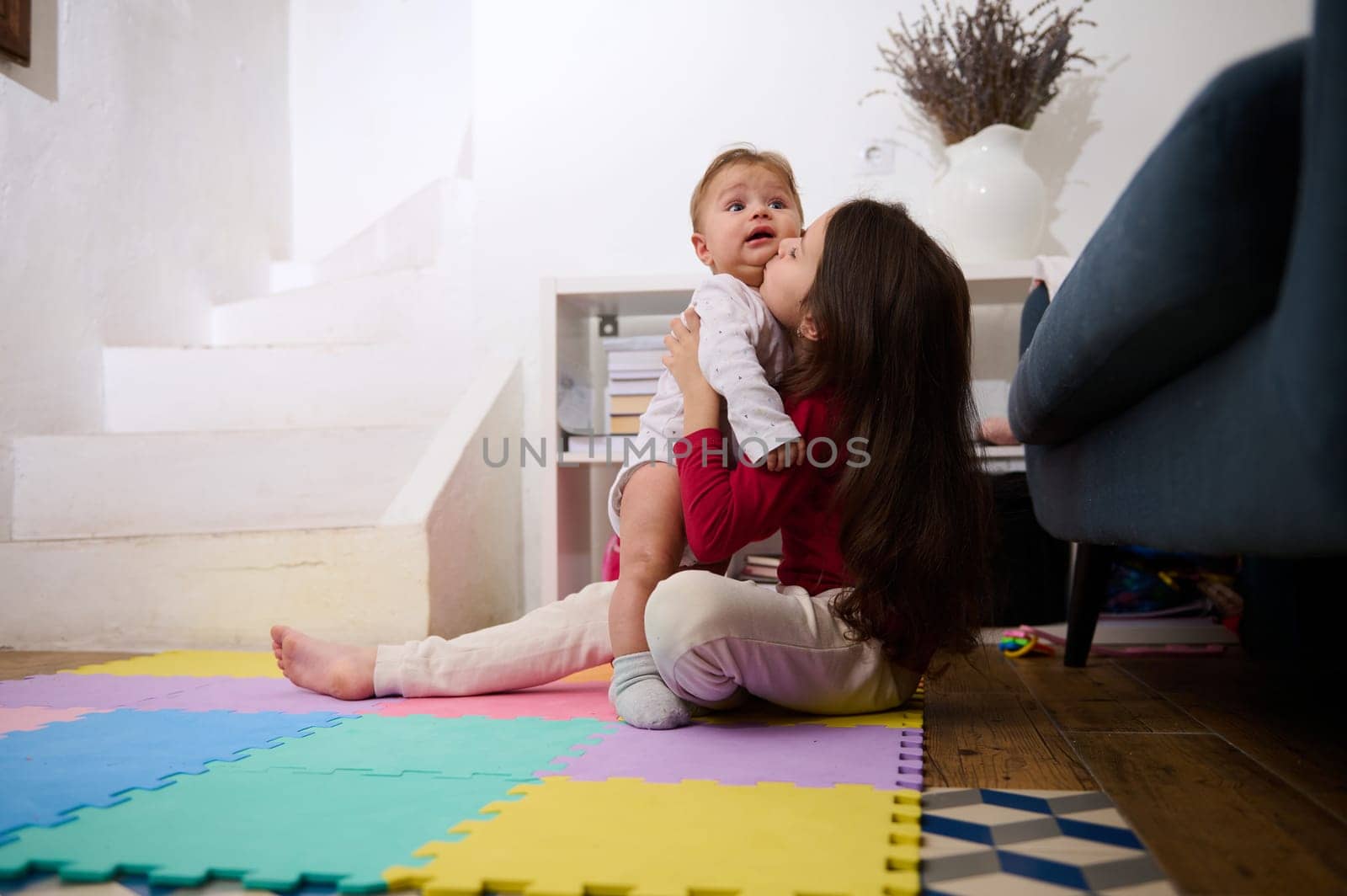 Adorable little child girl sitting on colorful puzzle carpet, holding and kissing her little brother, playing with a cute baby boy in light interior of cozy rural house. Family relationships concept by artgf