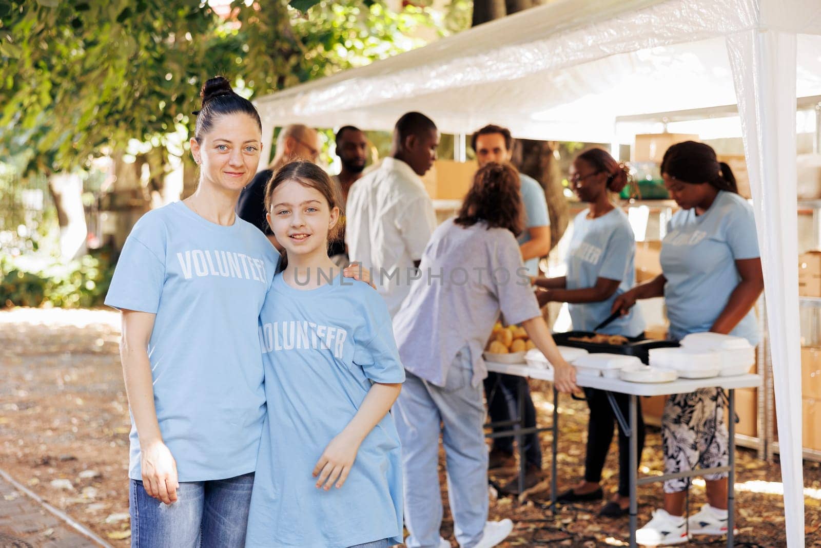Portrait shot of a mother and daughter, supporting hunger relief initiative at an outdoor food bank. Two caucasian females wearing blue t-shirts written volunteer, ready to help the homeless people.