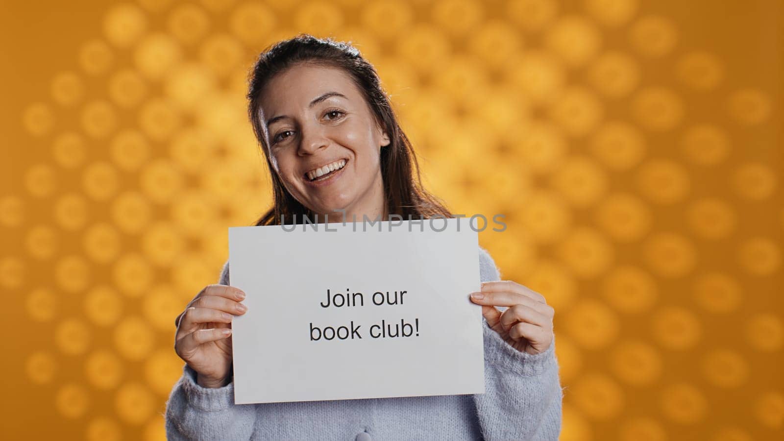Cheerful book club president holds message urging people to join them, talking about importance of lecture, studio background. Smiling woman inviting bookworms to enlist in her association, camera B