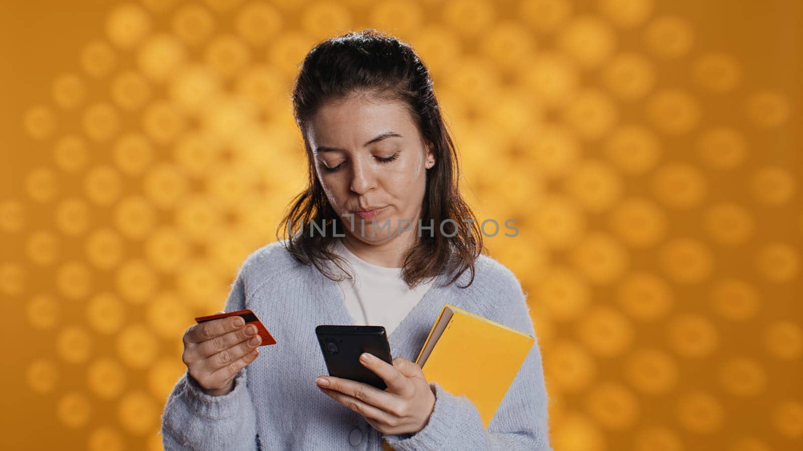 Woman holding books, purchasing more with phone, conveying joy of reading concept, studio background. Person buying novels online with credit card, showing appreciation for literature, camera B