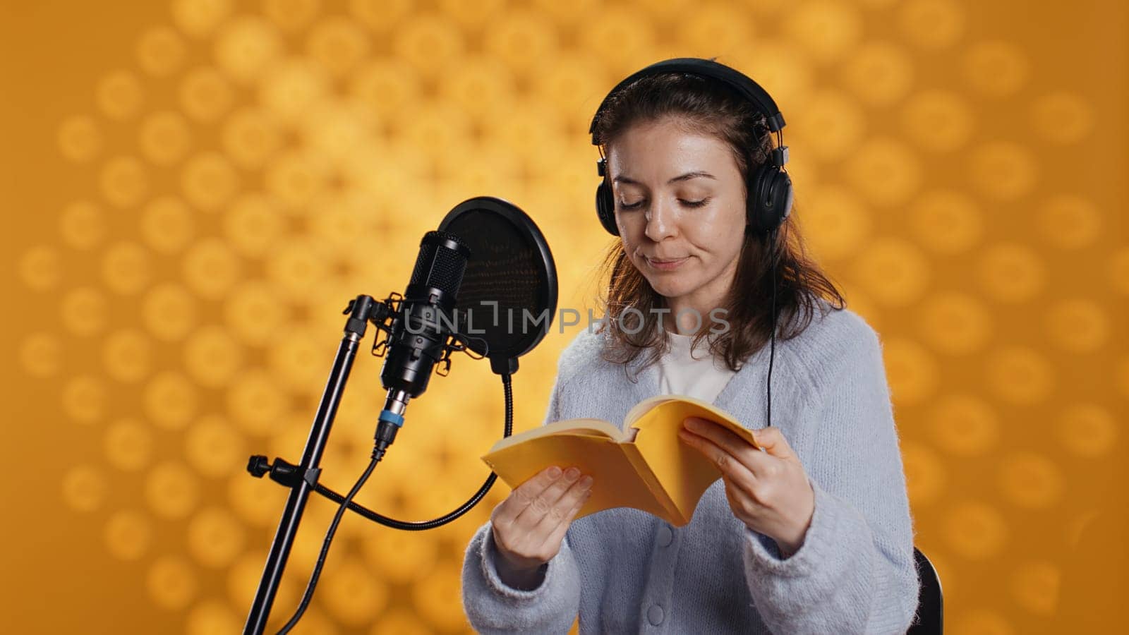 Influencer arriving in studio, putting headphones on, filming herself reading book with cellphone on tripod, creating audiobook. Woman doing vlog, recording novel using professional mic, camera B