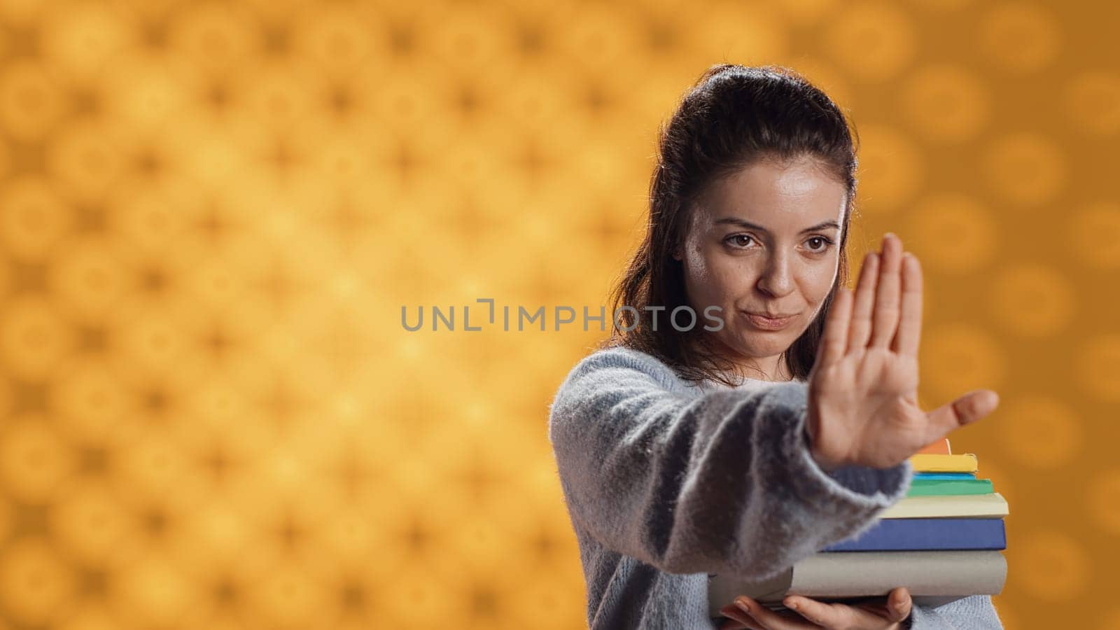 Portrait of stern woman holding stack of books doing stop sign gesturing, studio background. Student with pile of textbooks in arms used for academic learning doing halt hand gesture, camera A