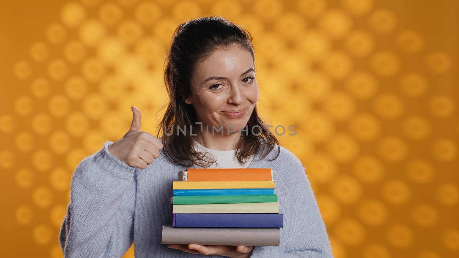 Portrait of happy woman with stack of books in hands showing thumbs up, studio background. Joyous bookworm holding pile of novels, feeling upbeat, doing positive hand gesturing, camera B