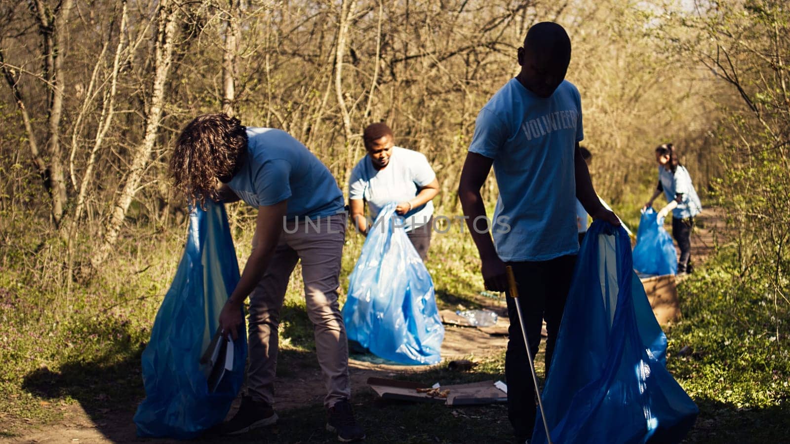 Diverse men volunteers pick up rubbish and plastic trash with tongs, working to combat illegal dumping and preserve natural forest environment. Activists volunteering for litter cleanup. Camera B.