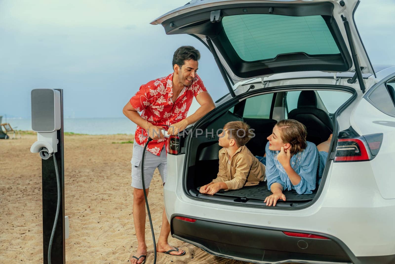 Family vacation trip traveling by the beach with electric car, lovely family sit on the trunk, charging EV car battery with green and sustainable energy. Family travel and eco-friendly car. Perpetual