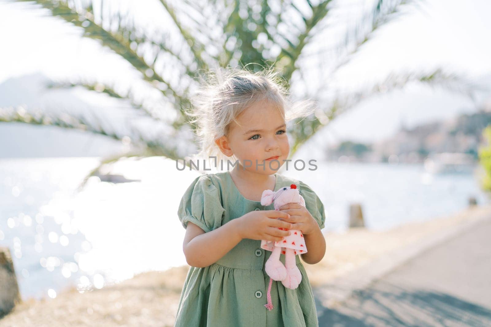 Little girl hugs a toy pink mouse while standing on the seashore near a green palm tree by Nadtochiy