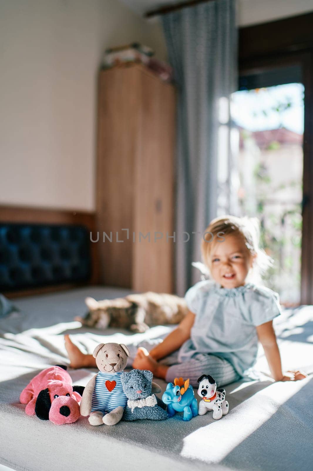 Teddy bear and soft toys sit in a row on the bed against the background of a little smiling girl by Nadtochiy