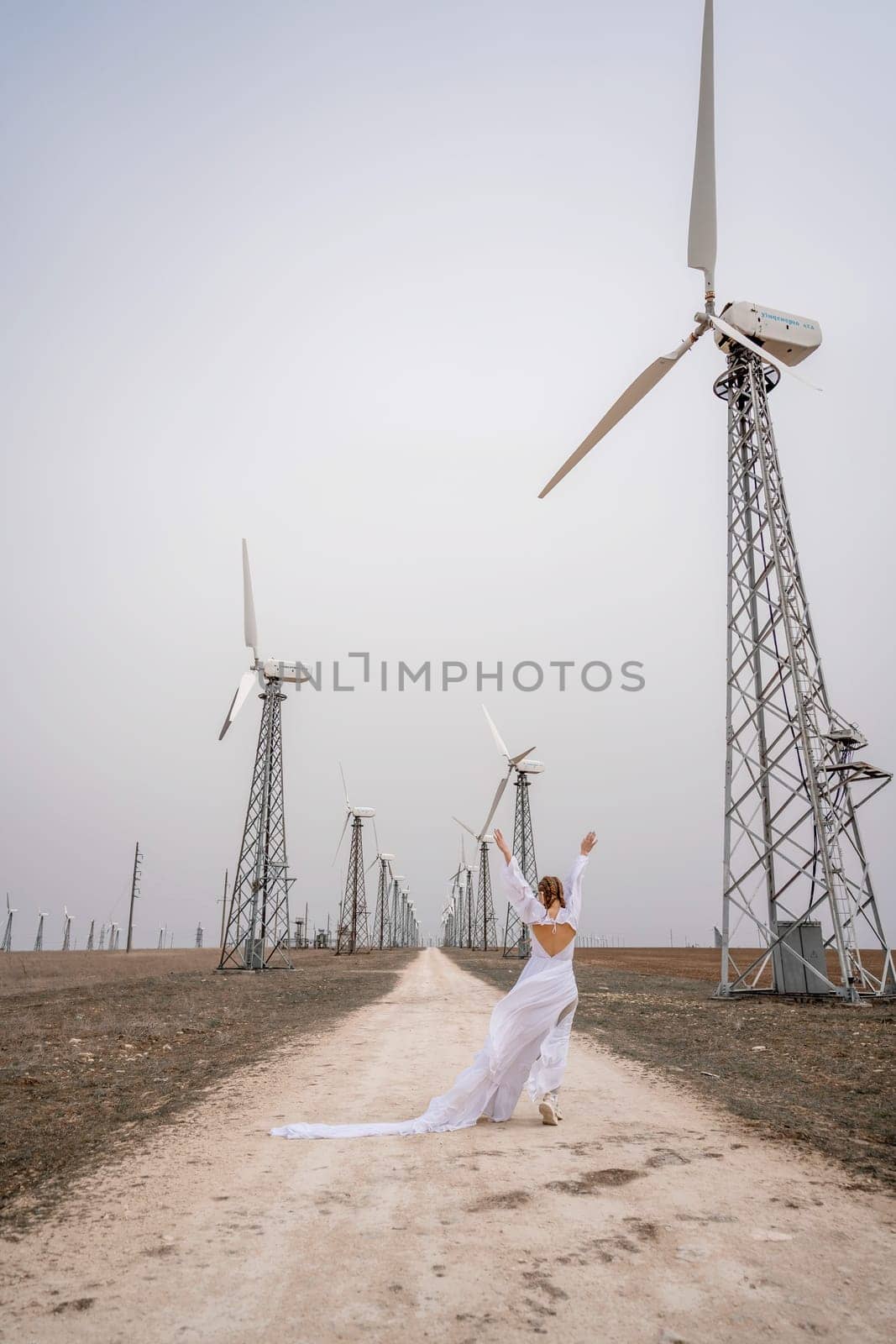 A woman in a white dress is walking down a dirt road in front of a row of wind turbines