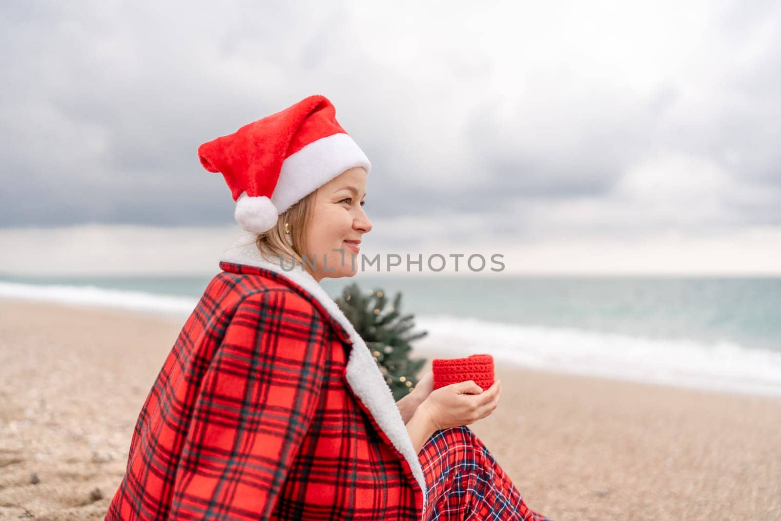Sea Lady in Santa hat plaid shirt with a red mug in her hands enjoys beach with Christmas tree. Coastal area. Christmas, New Year holidays concep.