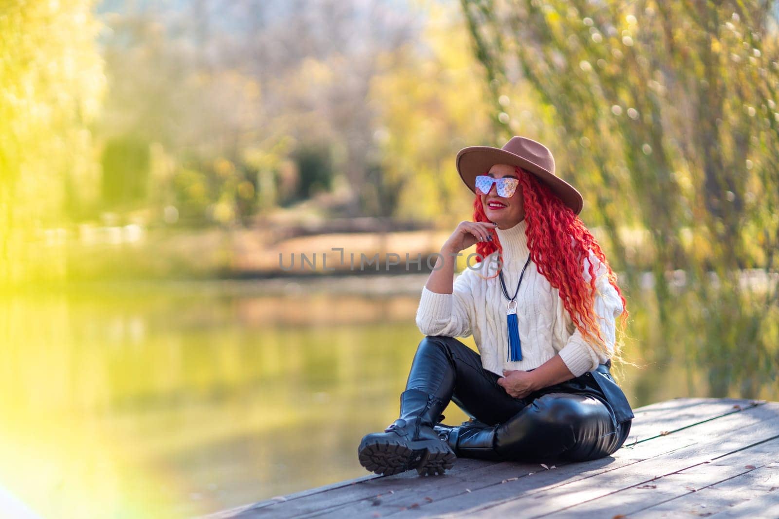 Autumn lake woman. In autumn, she sits by the pond on a wooden pier and admires nature with red hair and a hat. Tourism concept, weekend outside the city. by Matiunina