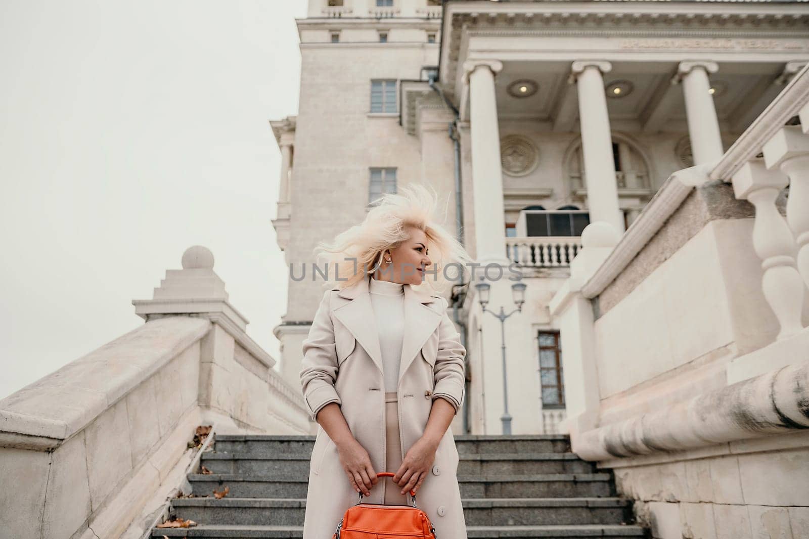 A woman wearing a white coat and blonde hair stands on a set of stairs in front of a large building. She is holding a red purse in her hand. Concept of elegance and sophistication. by Matiunina