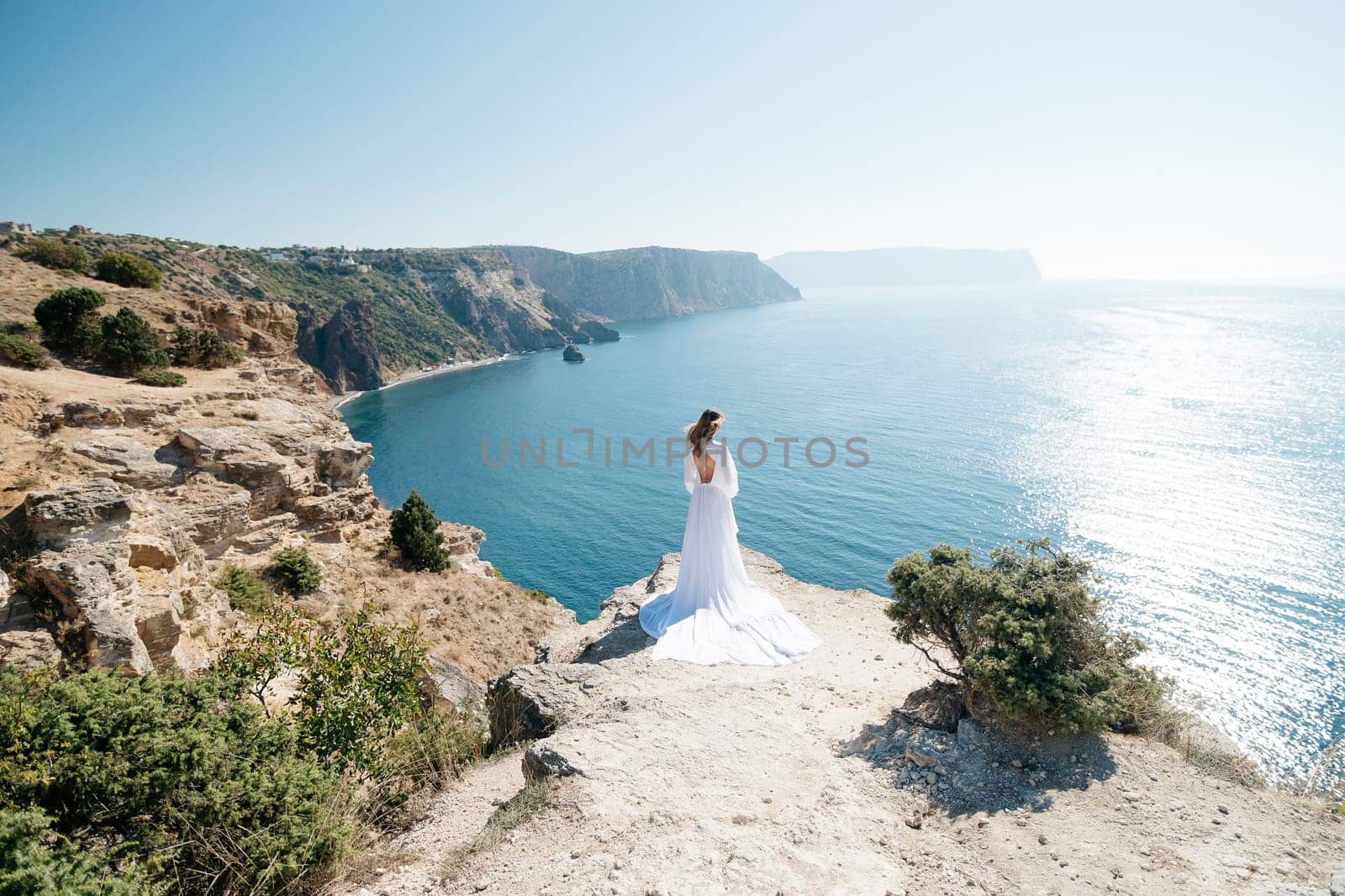 woman white dress stands on a rocky cliff overlooking the ocean. The scene is serene and peaceful, with the woman's dress billowing in the wind