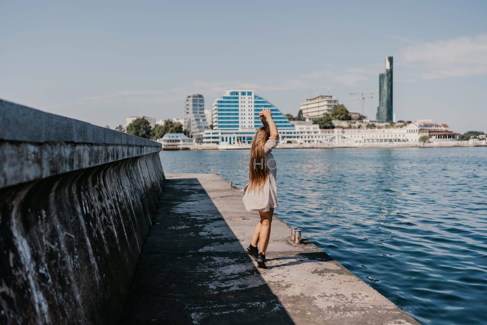 woman in a white dress is walking on a pier near the water. The scene is peaceful and serene, with the woman's long hair blowing in the wind. by Matiunina