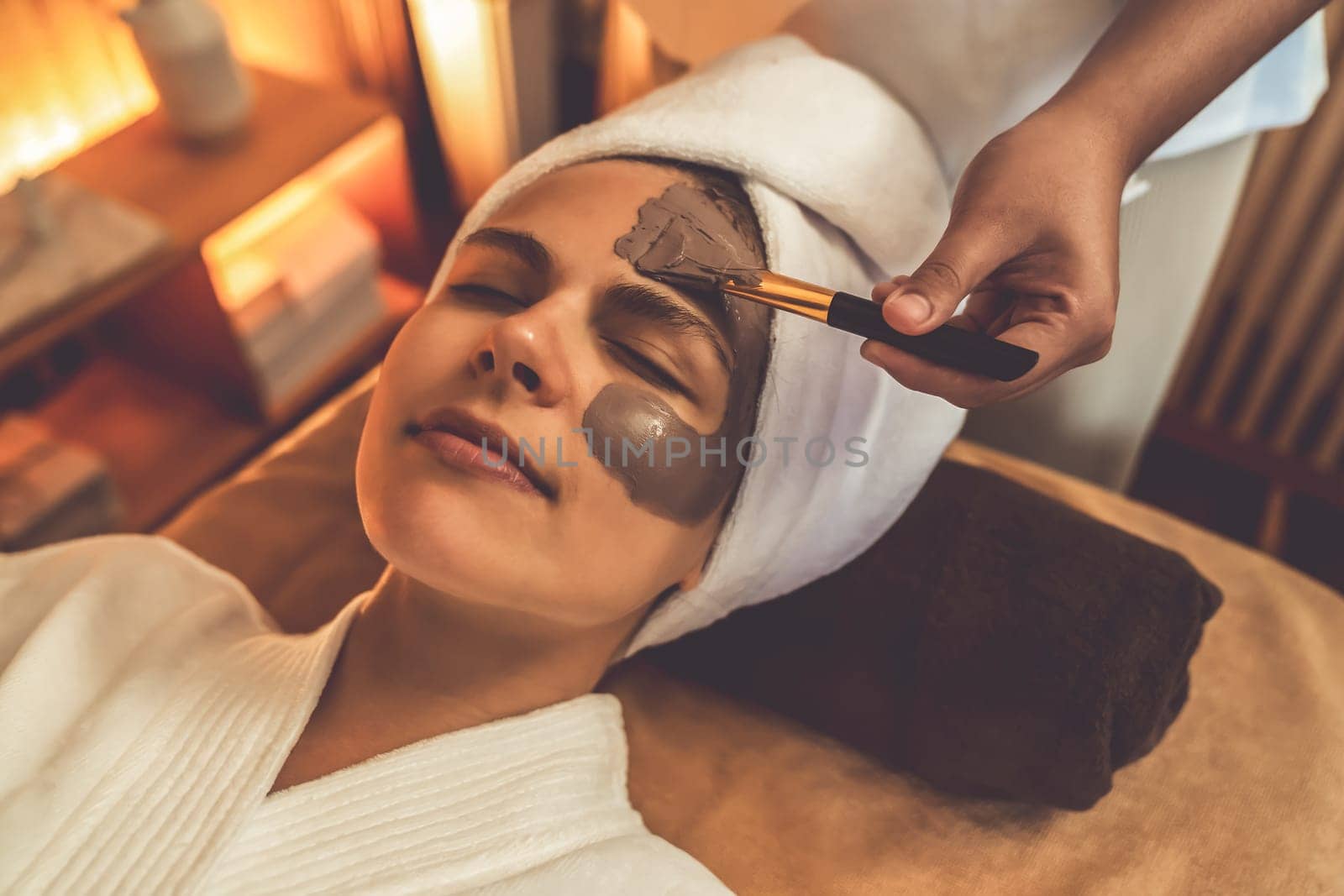 Serene ambiance of spa salon, woman customer indulges in rejuvenating with charcoal face cream massage with warm lighting candle. Facial skin treatment and beauty care concept. Quiescent