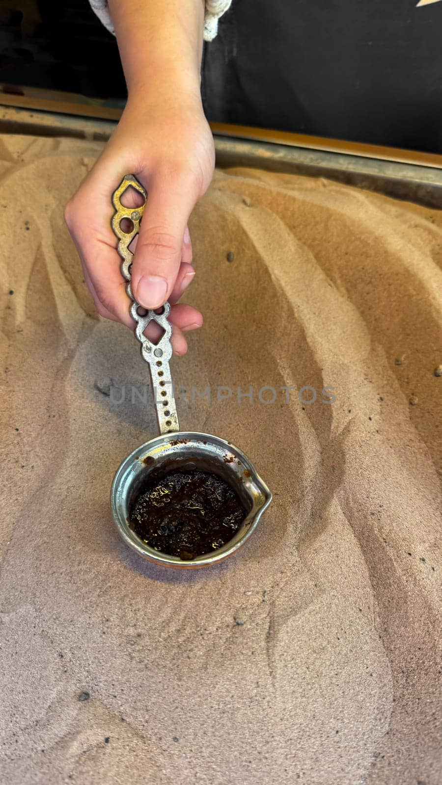 Hand holding traditional copper cezve with brewed Turkish coffee over sand, with coffee grounds visible and shadows playing on the sand. Turkish coffee making tradition. by Lunnica