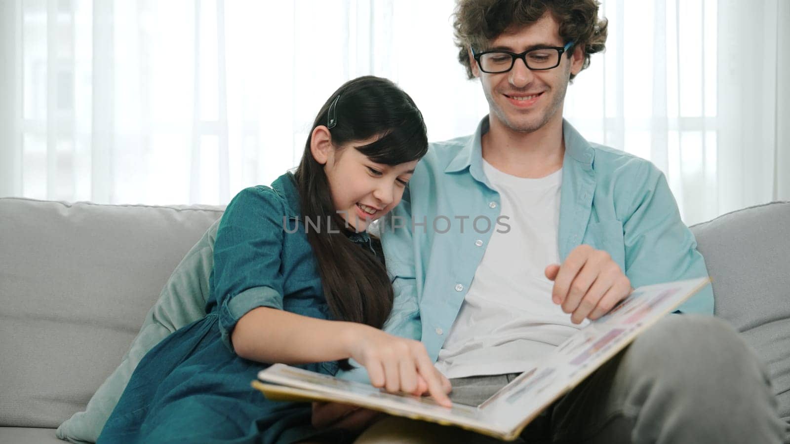 Happy daughter lean on father shoulder while listening story telling. Young asian schoolgirl looking ar dad while reading story book together and sitting at sofa. Family recreation concept. Pedagogy.