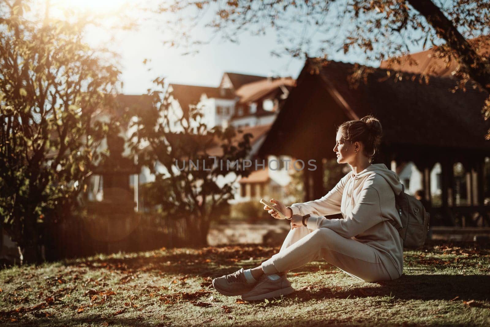 Young fashionable teenage girl with smartphone in park in autumn sitting at smiling. Trendy young woman in fall in park texting. Retouched, vibrant colors. Beautiful blonde teenage girl wearing casual modern autumn outfit sitting in park in autumn. Retouched, vibrant colors, brownish tones.