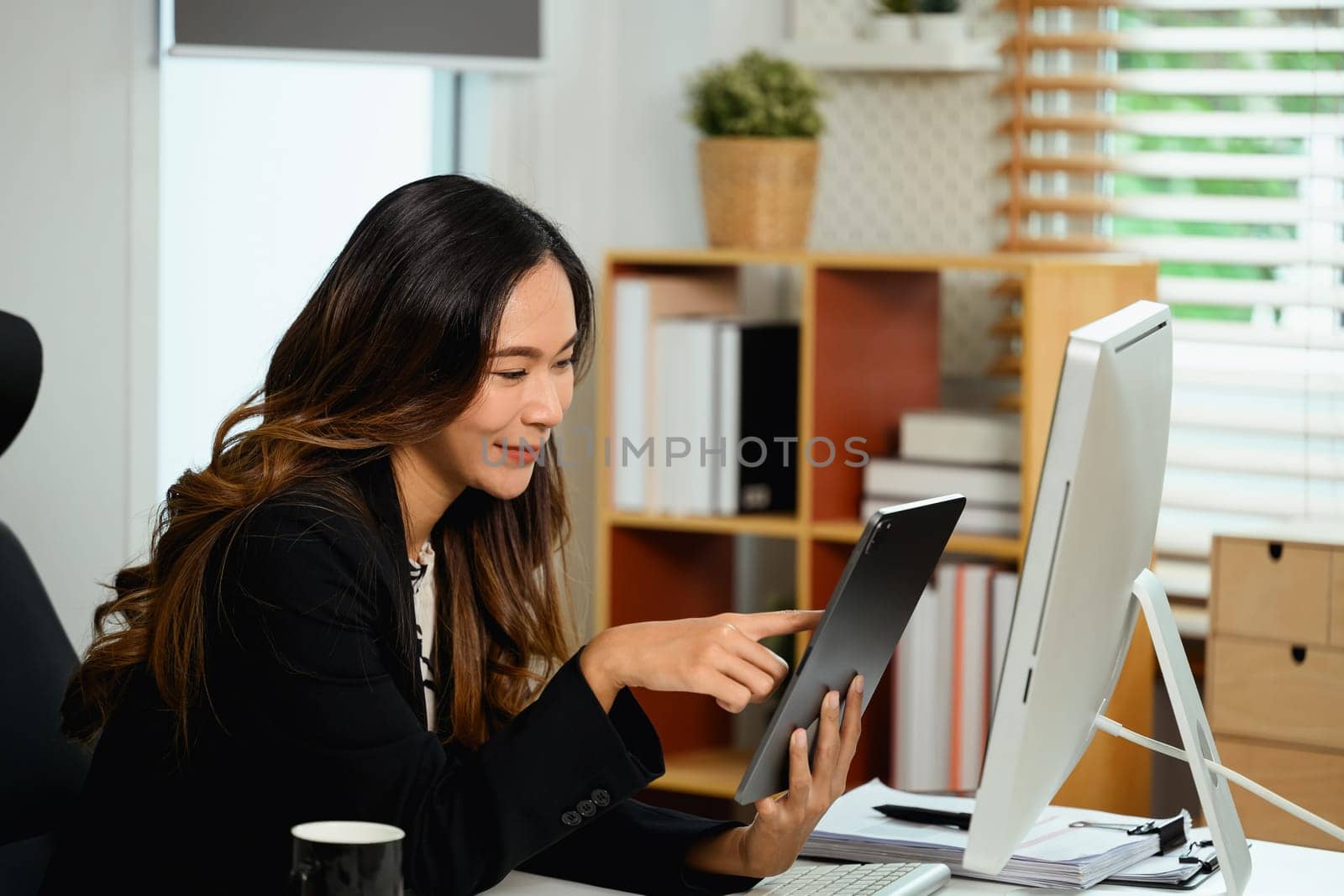 Cheerful young businesswoman sitting at desk and using digital tablet by prathanchorruangsak