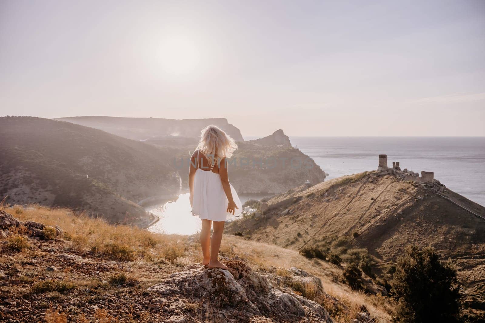 A woman stands on a hill overlooking a body of water. The sky is clear and the sun is shining brightly. The woman is enjoying the view and the peaceful atmosphere