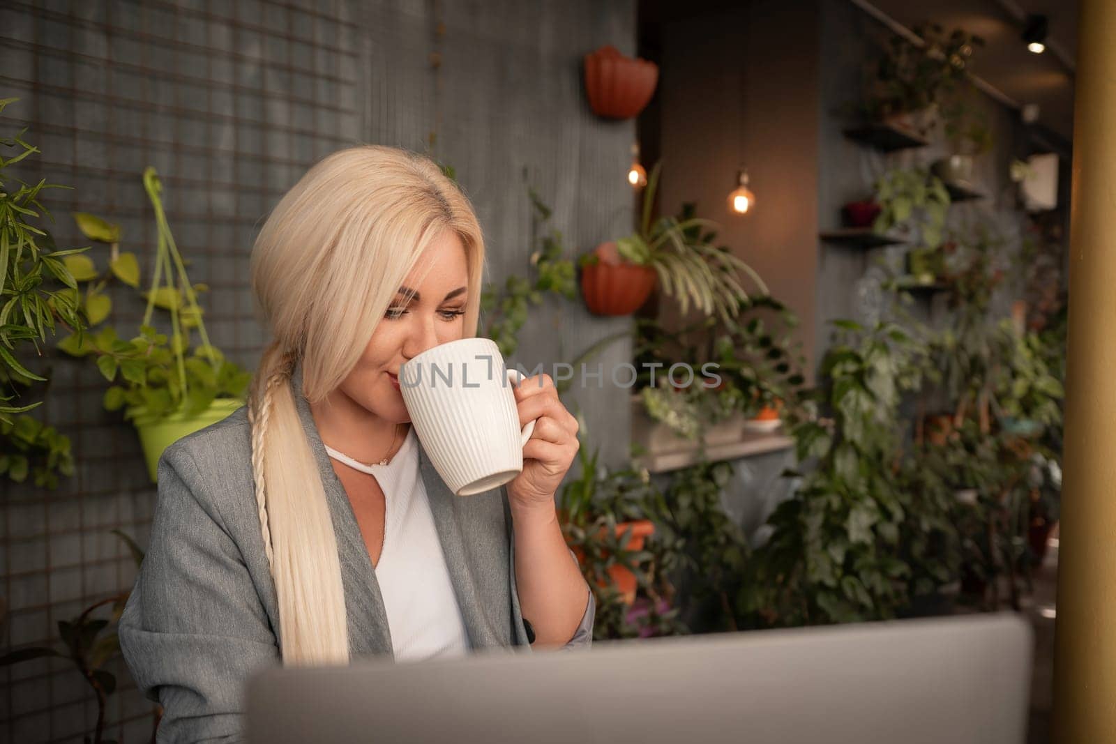 A blonde woman is sitting at a table with a laptop and a white coffee cup. She is drinking coffee while working on her laptop
