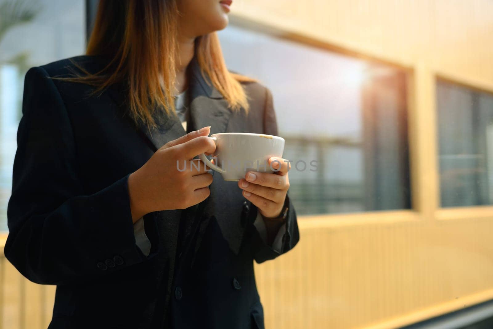 Businesswoman in black suit looking away while standing with coffee cup in modern office.