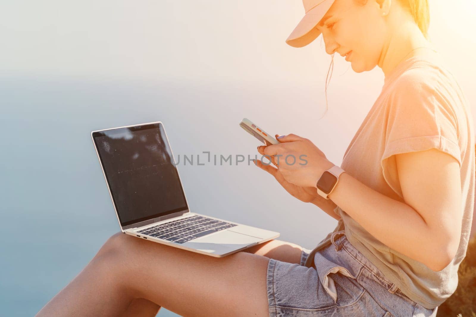 Digital nomad, woman in the hat, a business woman with a laptop sits on the rocks by the sea during sunset, makes a business transaction online from a distance. Freelance, remote work on vacation.