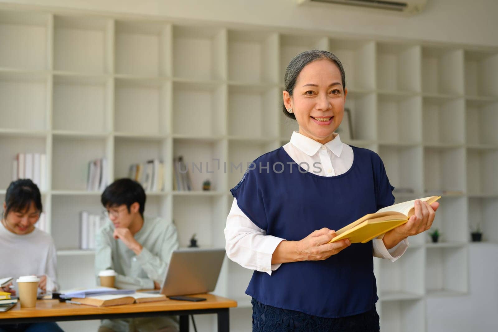 Portrait of friendly smiling mature professor holding book standing in class.
