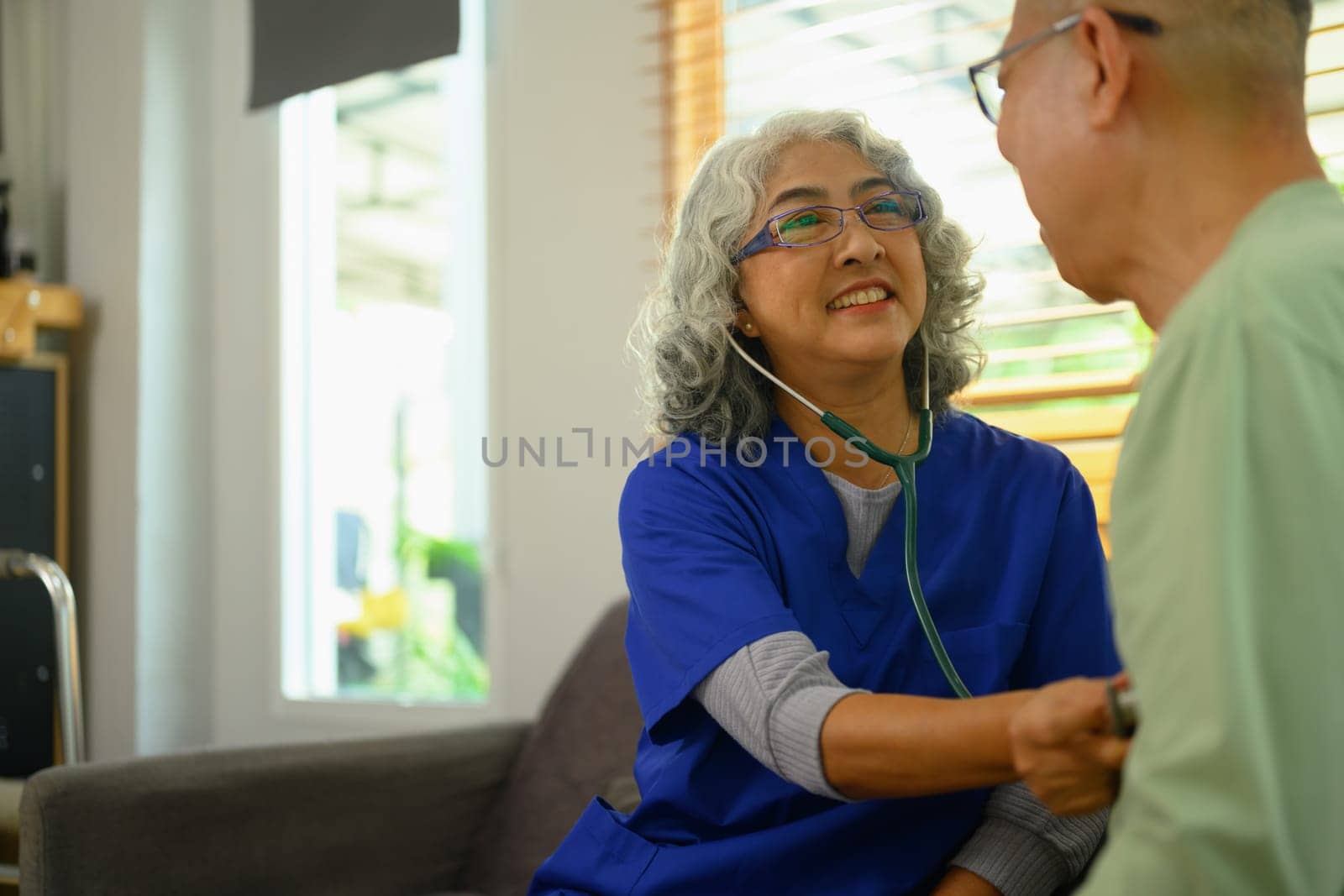 Smiling female doctor using stethoscope examining senior male during home care visit. Elderly healthcare concept.