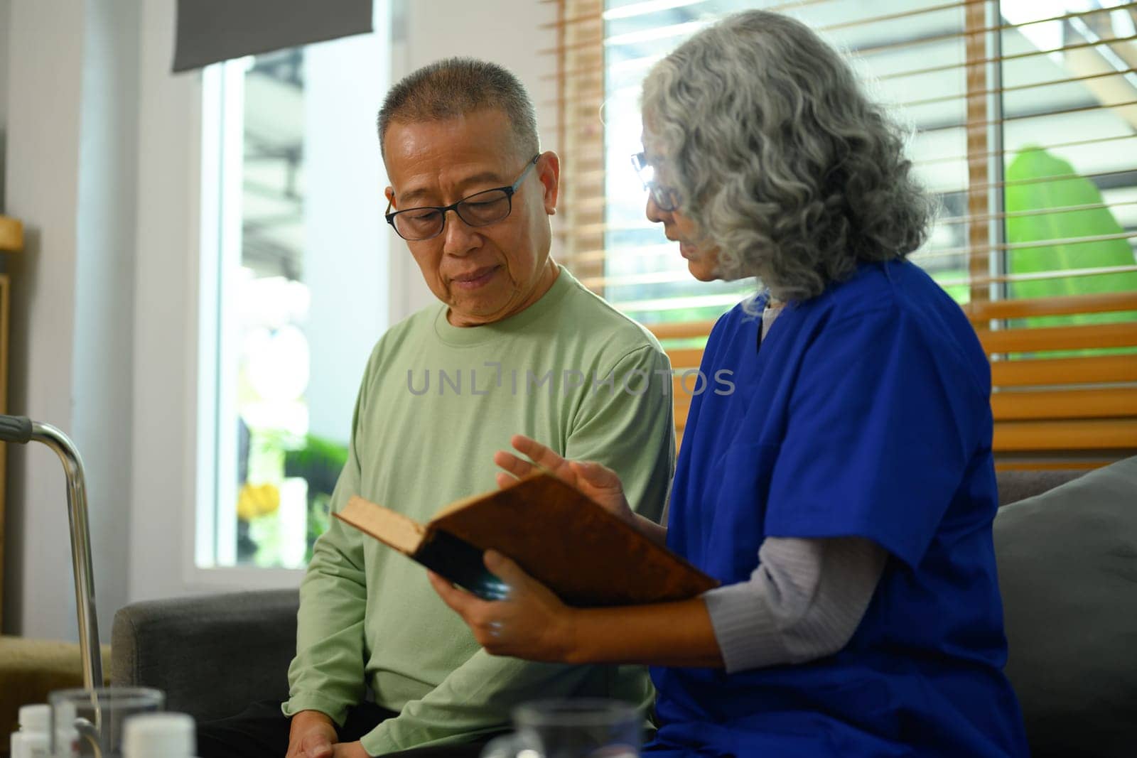 Nurse reading book to elderly man on sofa at nursing home. Volunteer and home health care service concept.