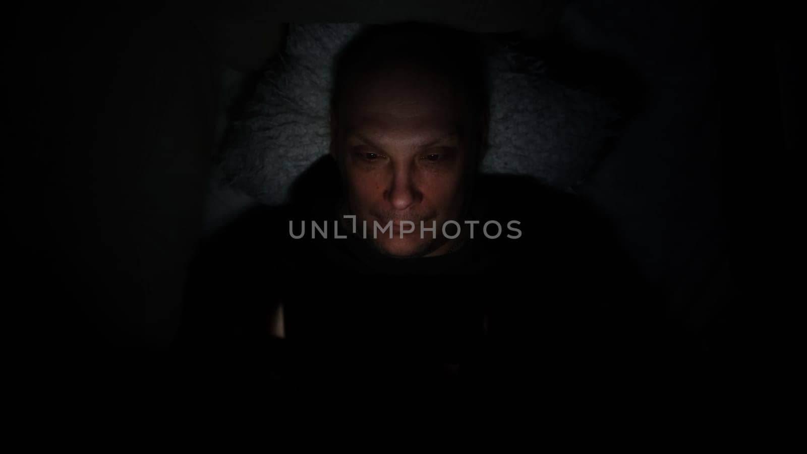 Lonely caucasian middle ages man lie on bed in bedroom and using phone at night. Face illuminated from smartphone display. Surfing social network before sleep.