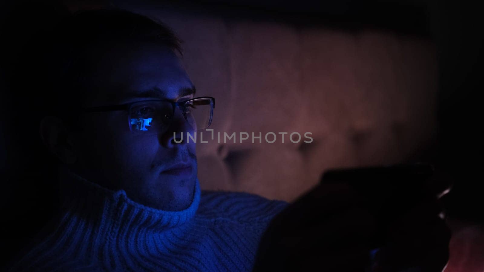 Lonely man in glasses sitting on bed in bedroom and using phone at night. Face illuminated from smartphone display. Surfing social network before sleep.