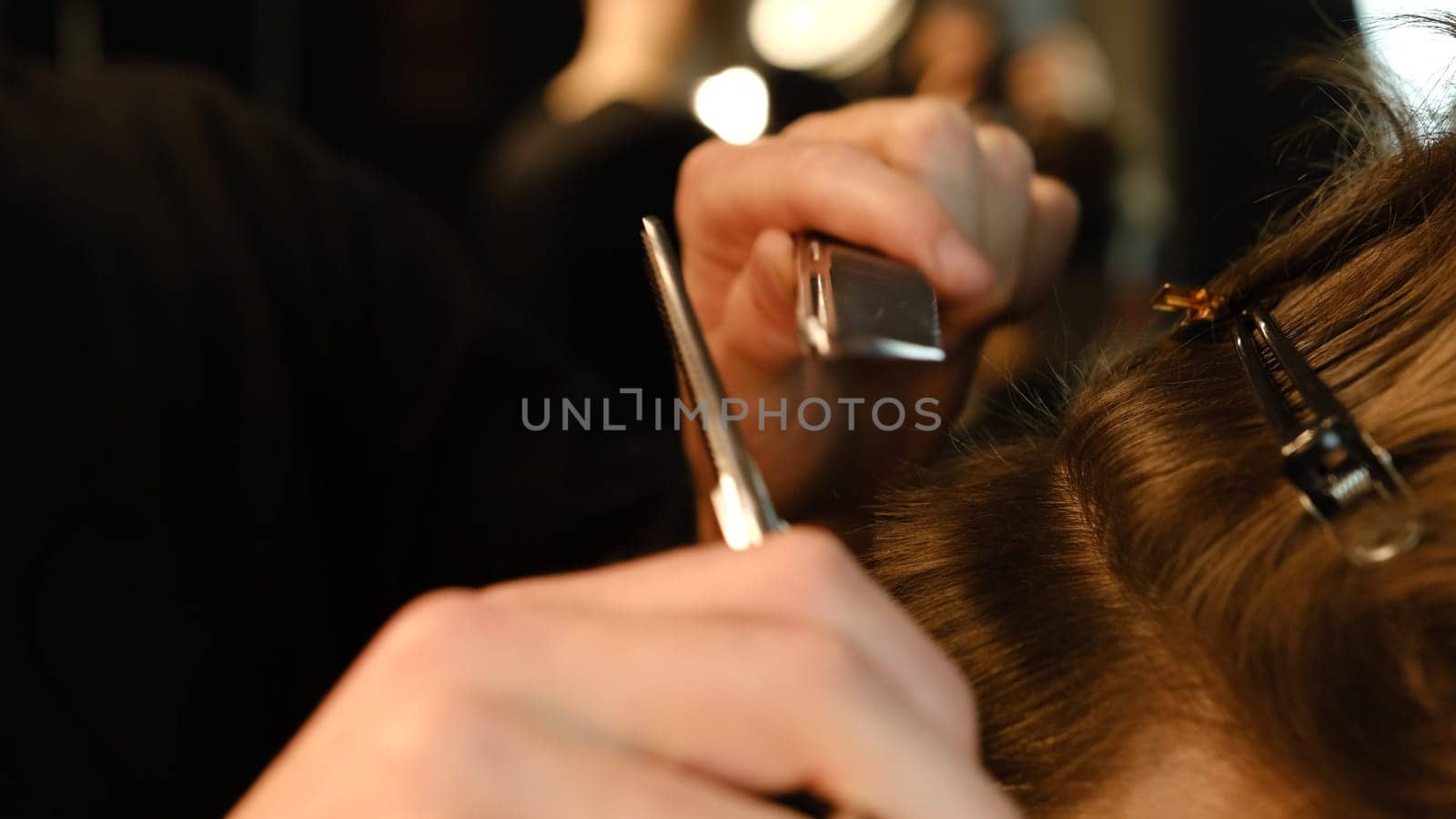 Close up Barber use Thinning scissors and metal comb on brown wavy hair of young man. Hairdresser service in a modern barbershop in a dark key lightning with warm light side view.