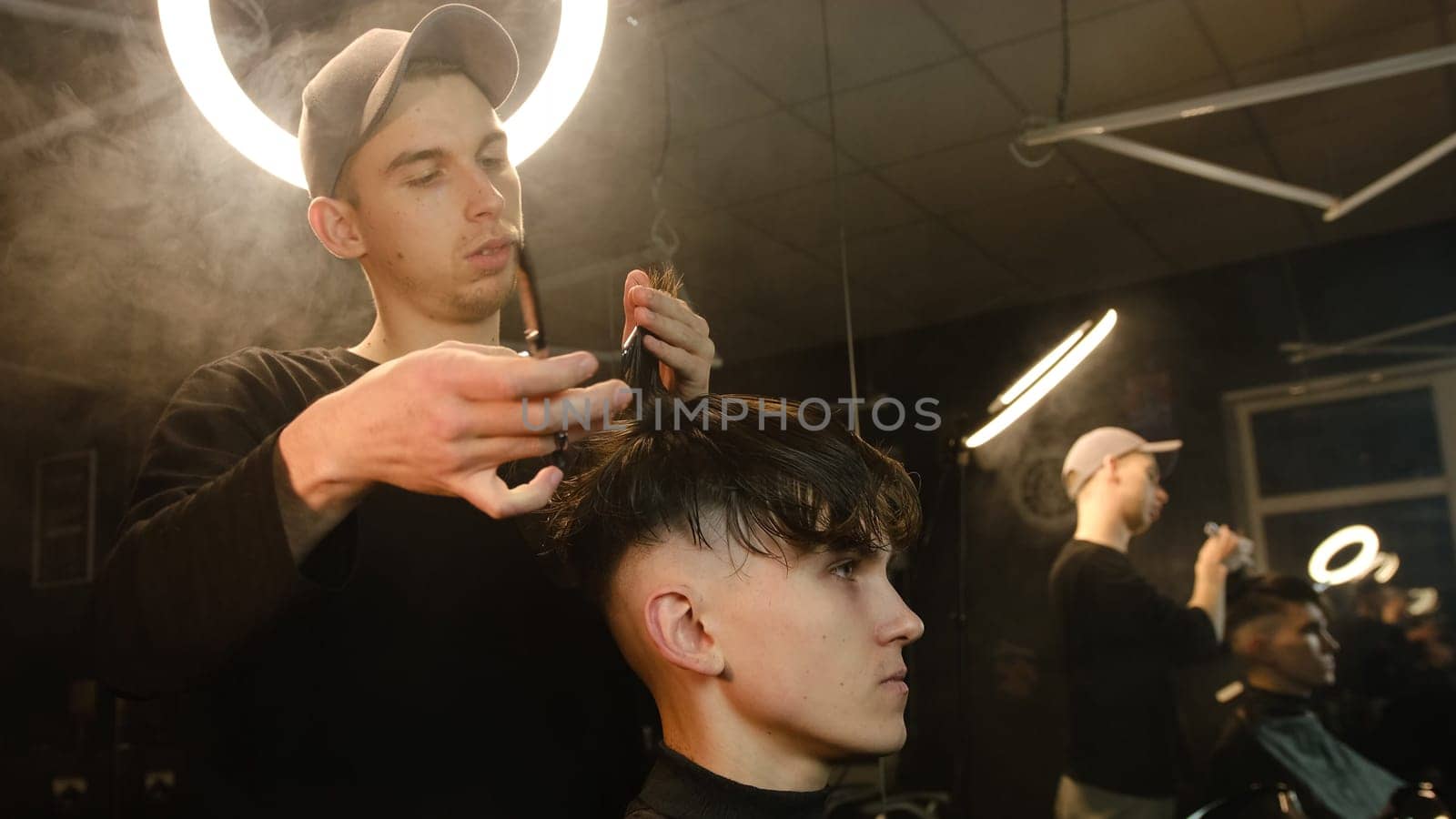Barber master hairdresser does hairstyle and style with scissors and comb. Concept Barbershop. side view in dark key lightning with warm light and smoke.
