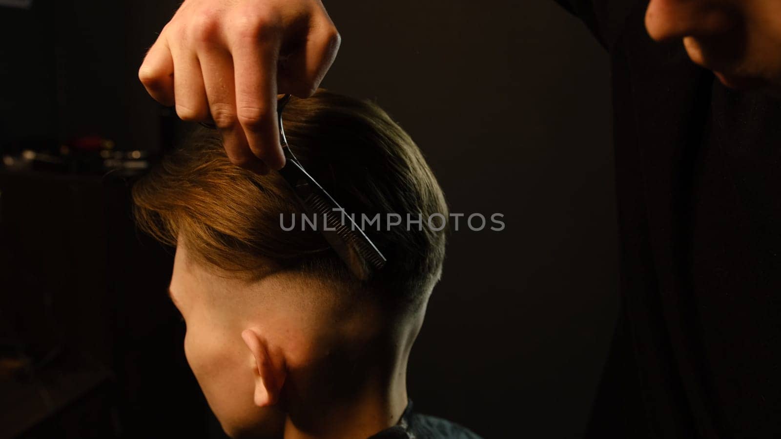 Barber use Thinning scissors and metal comb on brown wavy hair of young man. Hairdresser service in a modern barbershop in a dark key lightning with warm light.