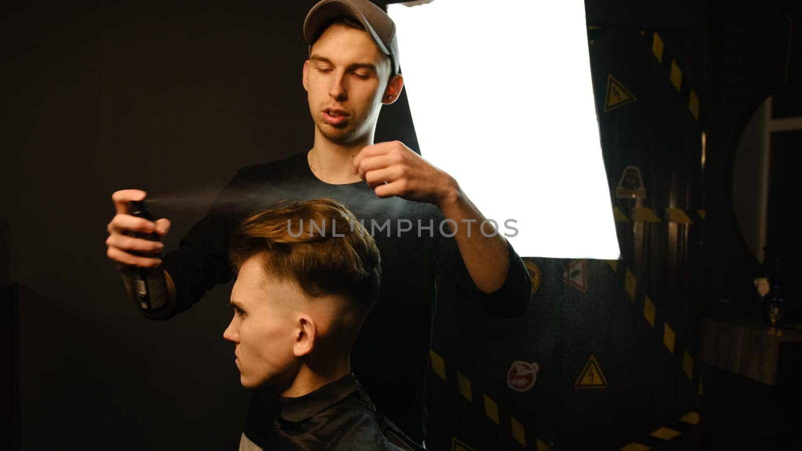 Barber makes hair styling with hair spray after haircut at the barber shop. Young handsome Caucasian man getting a haircut in a modern hairsalon from professional hairdresser.