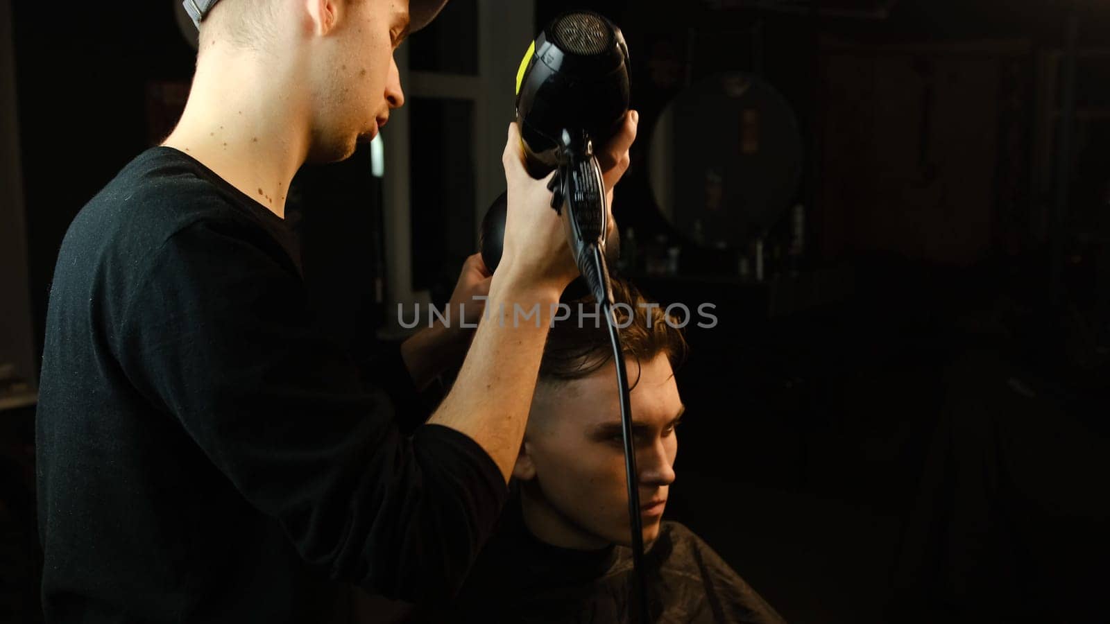 Young barber drying hair of customer with hair dryer in barber shop in a dark key lightning with warm light and correct hair with hand. Hairdresser for man.